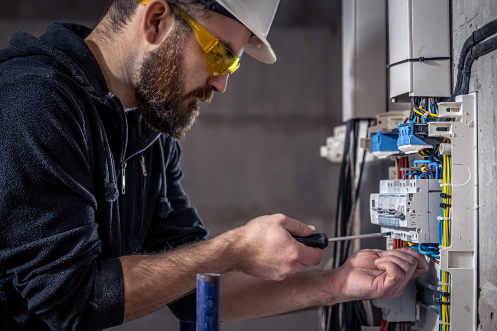 A male electrician works in a switchboard with an electrical connecting cable, connects the equipment with tools.