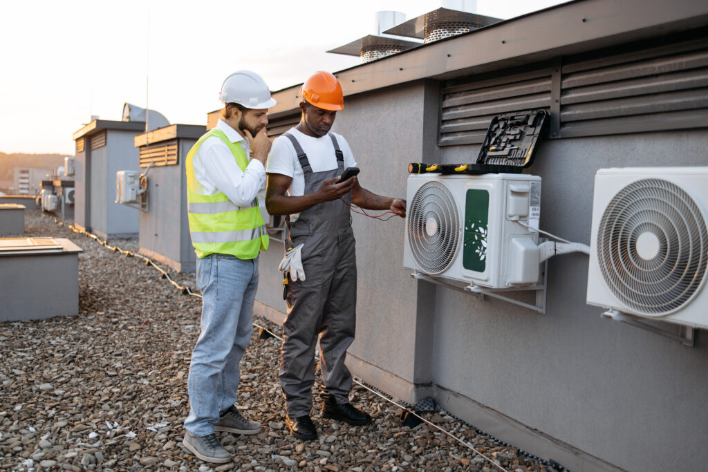 Team of two multiracial technicians checking voltage with modern multimeter and discussing repairing of cooling system. Competent male inspectors with toolbox looking at air conditioner outdoors.